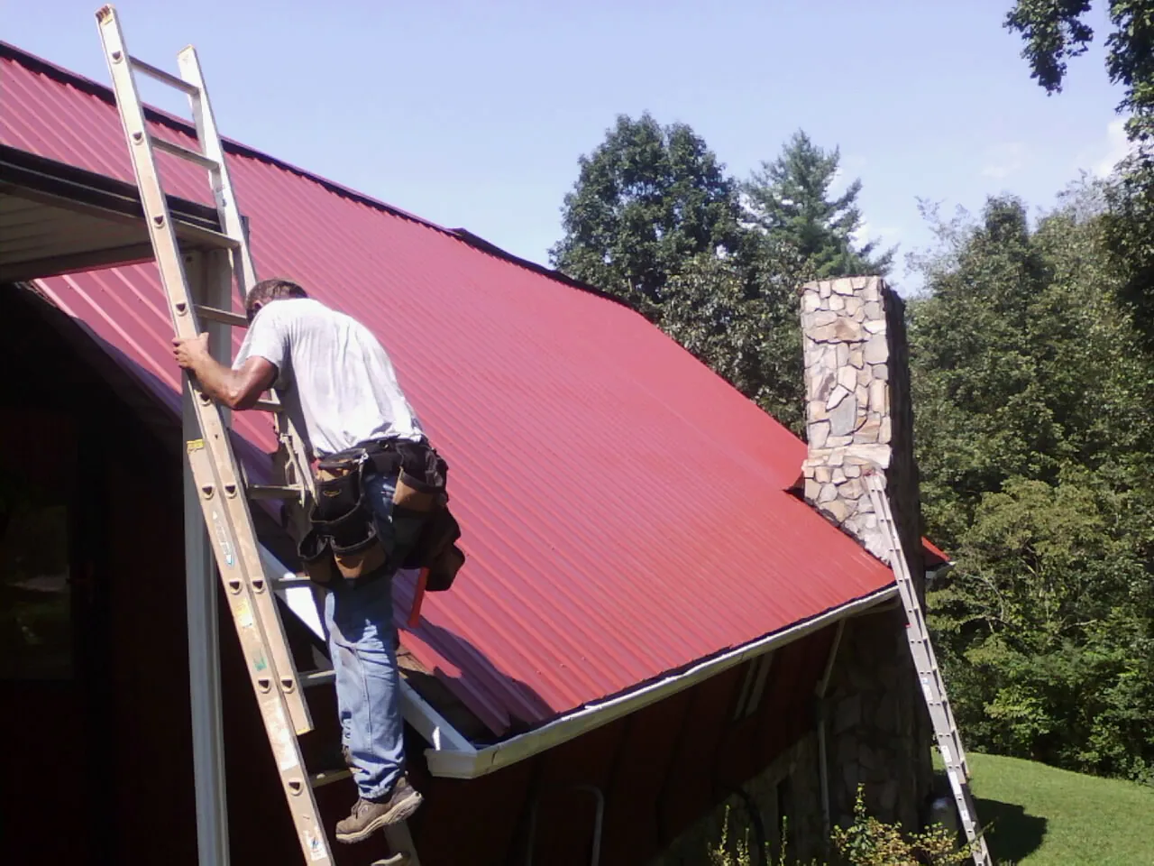 Roofer on a ladder beside a red metal roof and stone chimney — Soffit & Fascia Repair by Roofing Brothers in Agoura Hills